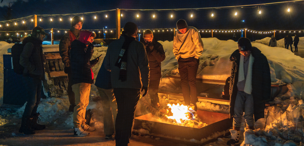 Students standing around a fire with a lighted ice rink in the background.