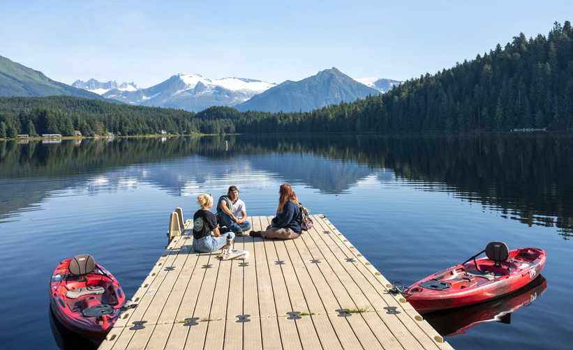 Three students sitting on a dock at UAS with mountains in the distance.